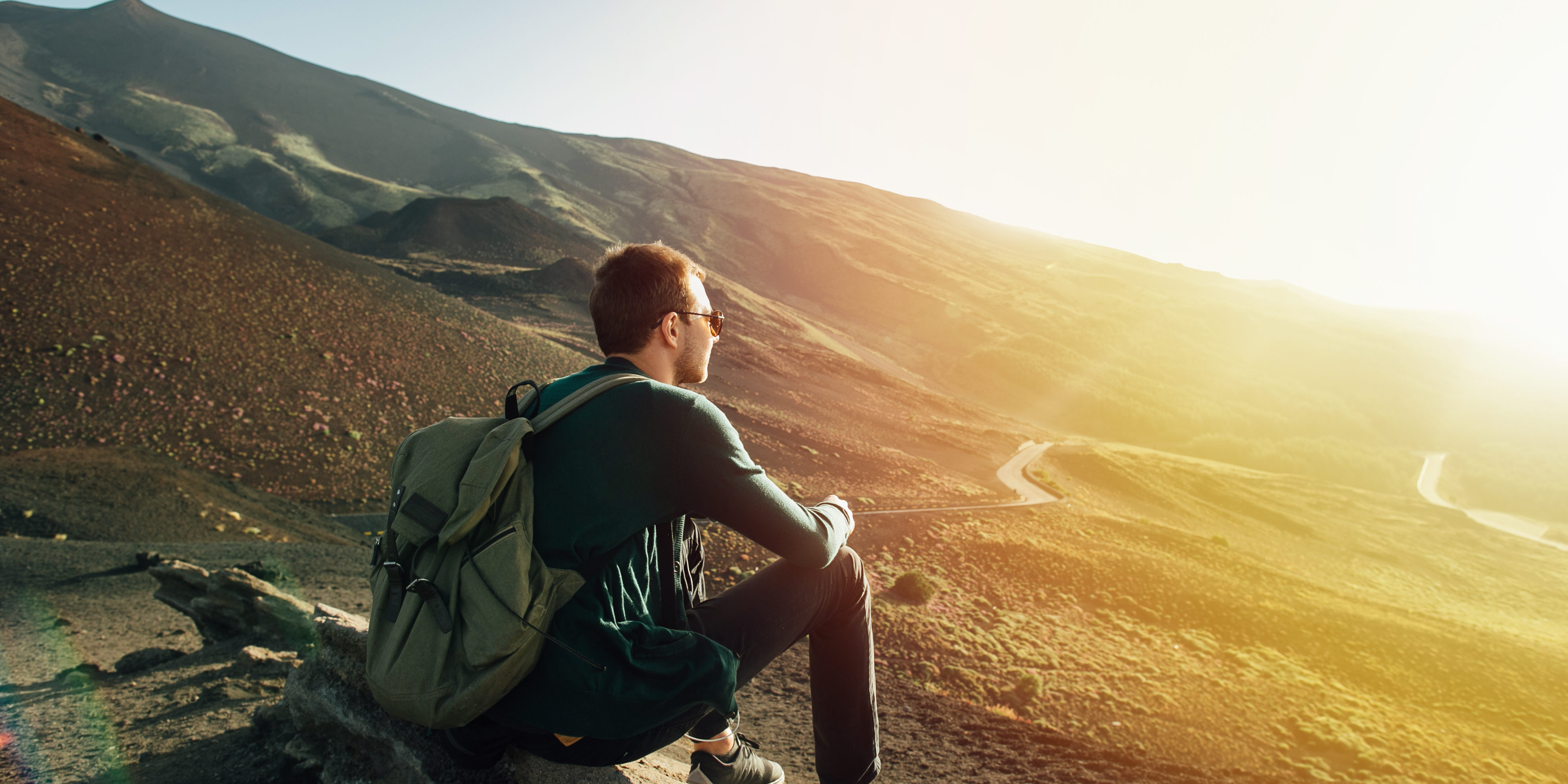 Man with rucksack sitting on rock at sunset on background of volcano Etna mountain in Sicily